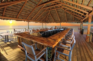 Eine Holzterrasse mit Barbereich, Sitzgelegenheiten im Freien und Blick auf den Sonnenuntergang über dem Meer.