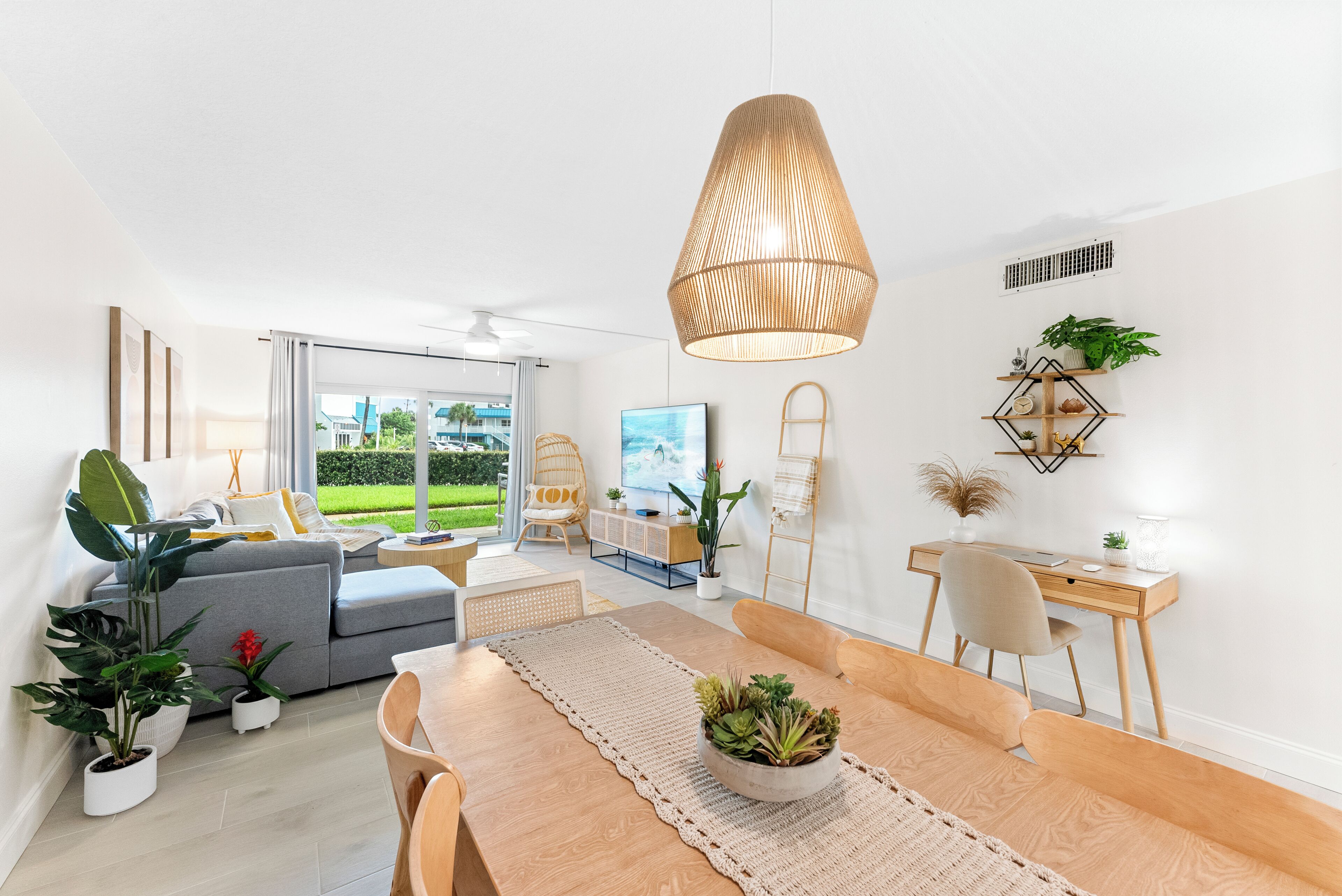 A modern living room with a dining area, a large window with a view of greenery, a ceiling fan, and a pendant light.