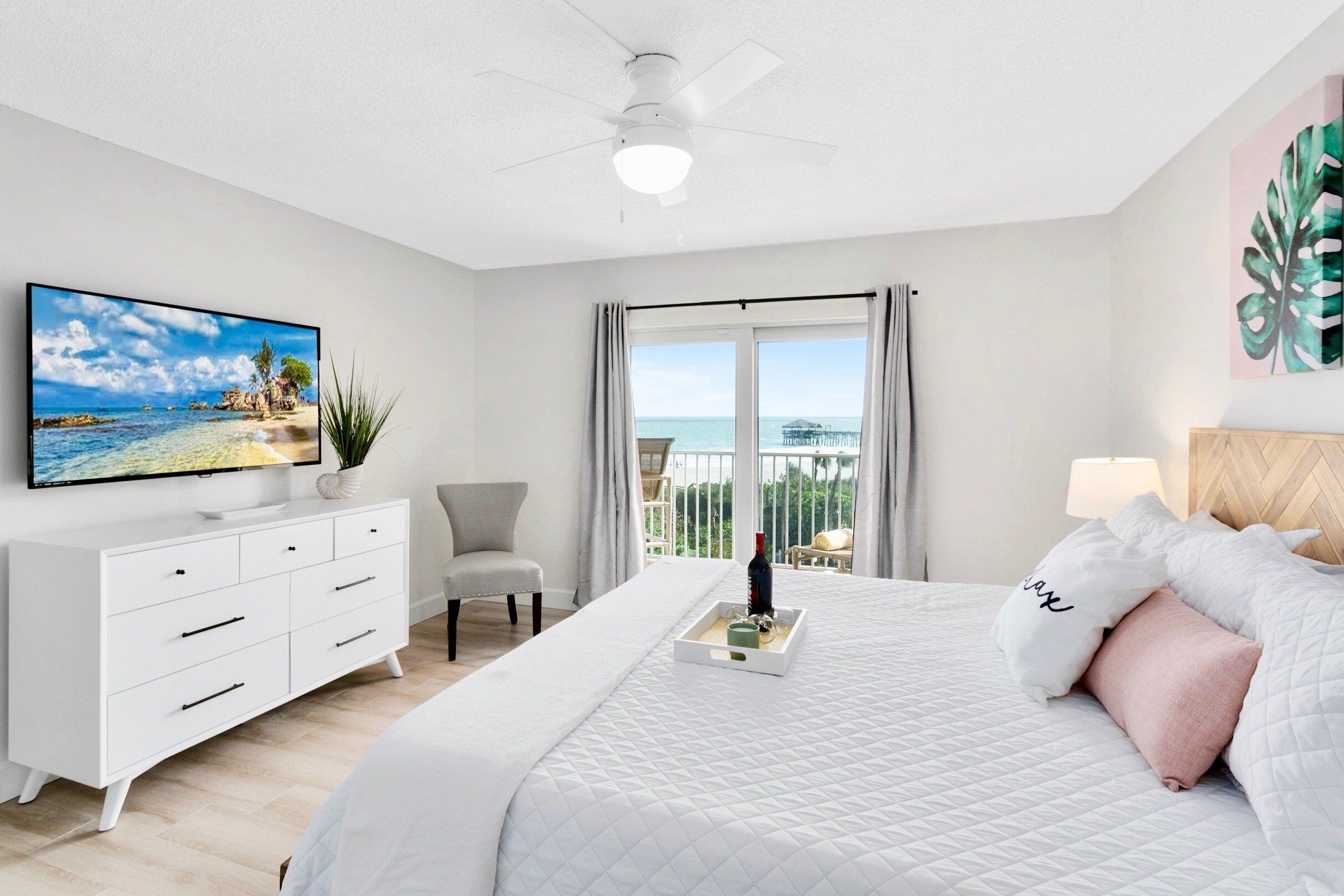 A bedroom with a bed, a TV, a dresser, a chair, and a balcony with a view of the sea.