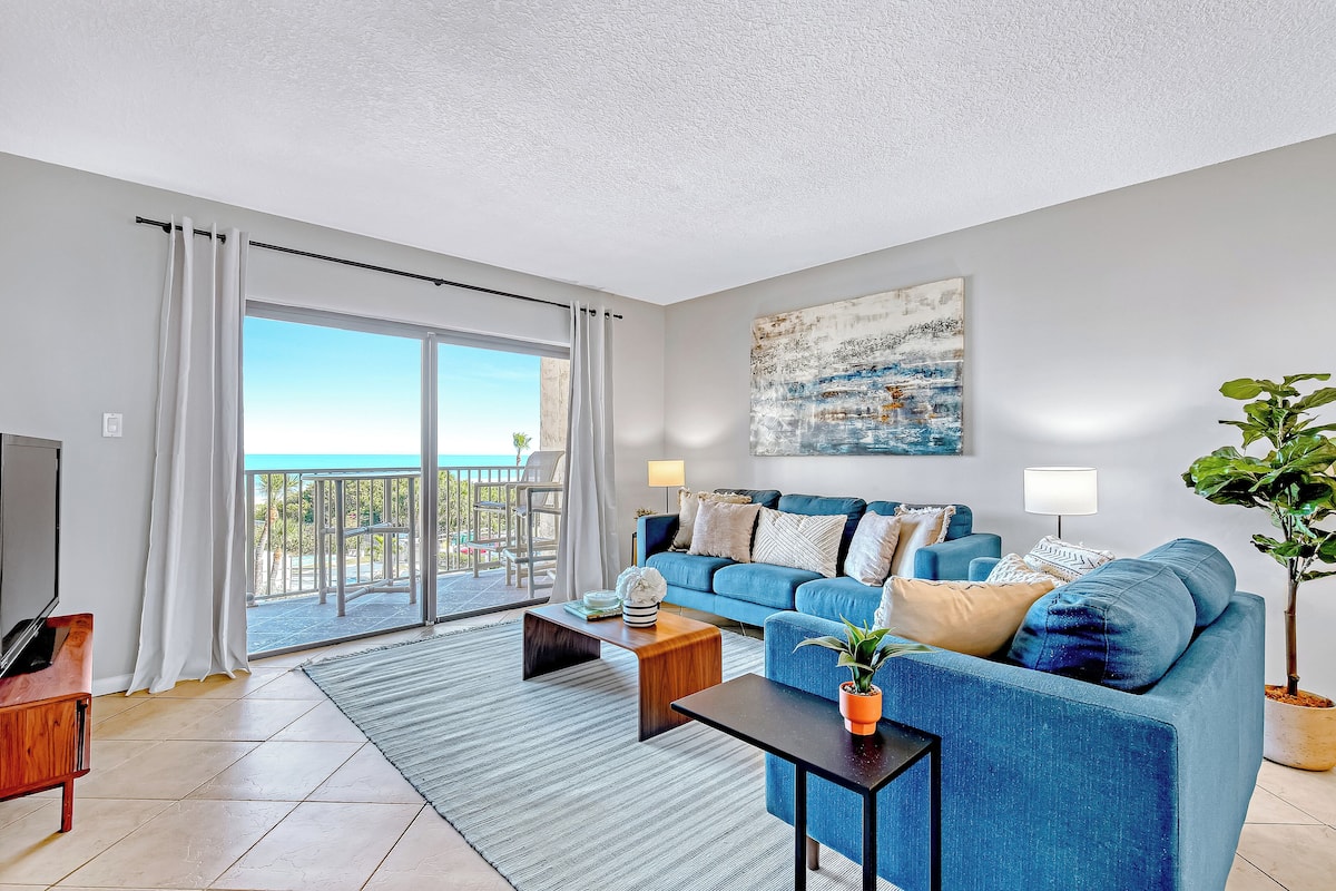 A modern living room with a blue sofa, a coffee table, a TV, and a balcony with a view of the sea.