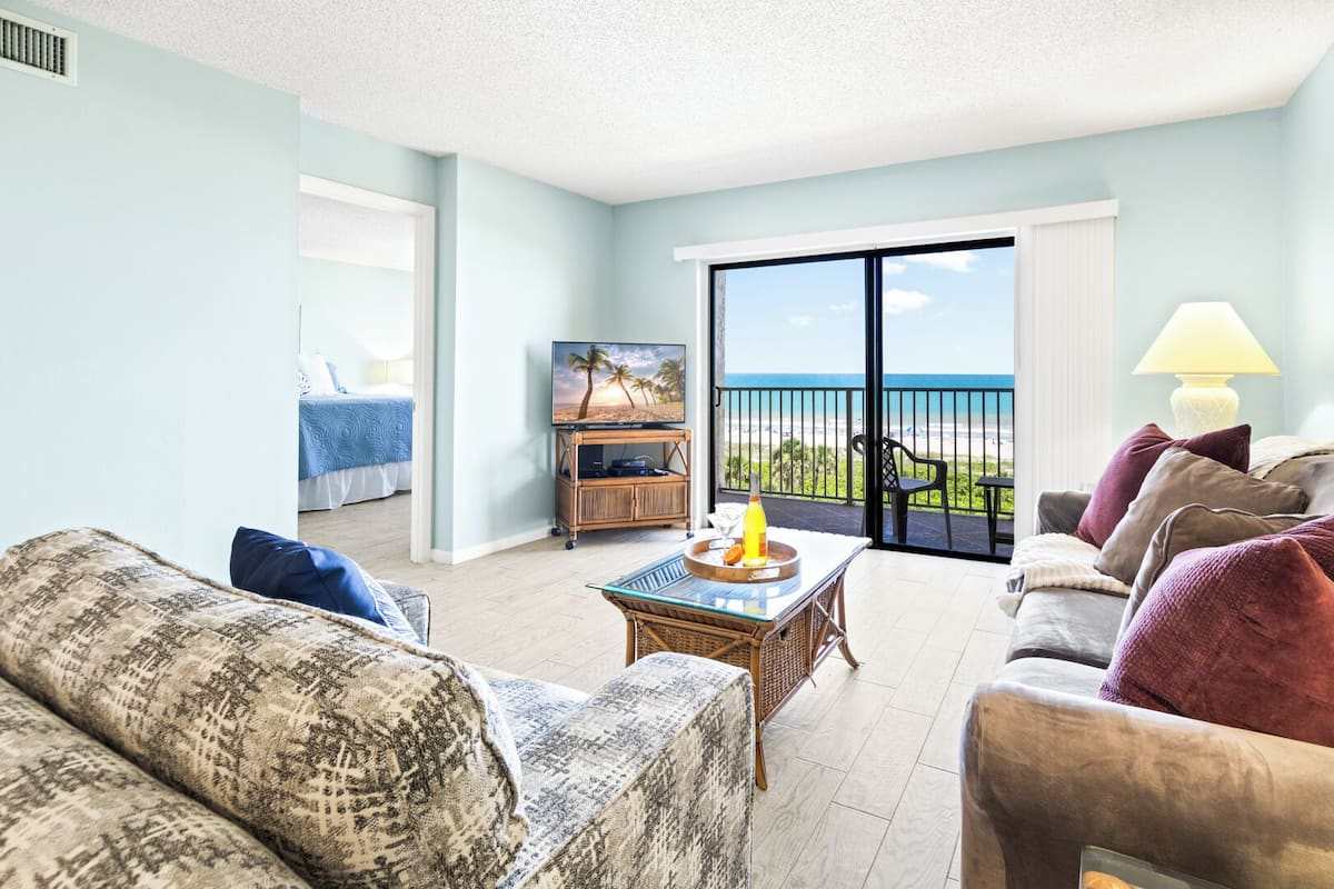 A living room with a sofa, a coffee table with a bottle and glass, a TV on a stand, and a balcony with a view of the beach.