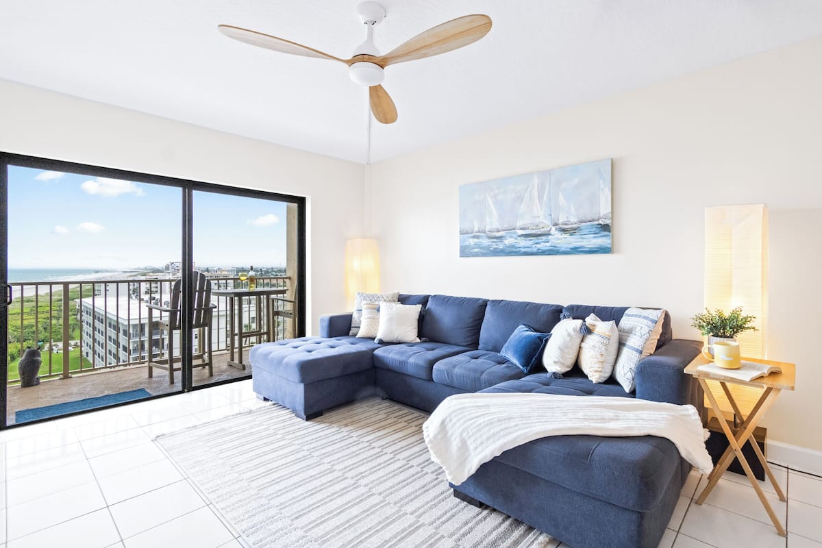 A modern living room with a sectional sofa, a ceiling fan, a painting of sailboats, and a view of the city through large windows.
