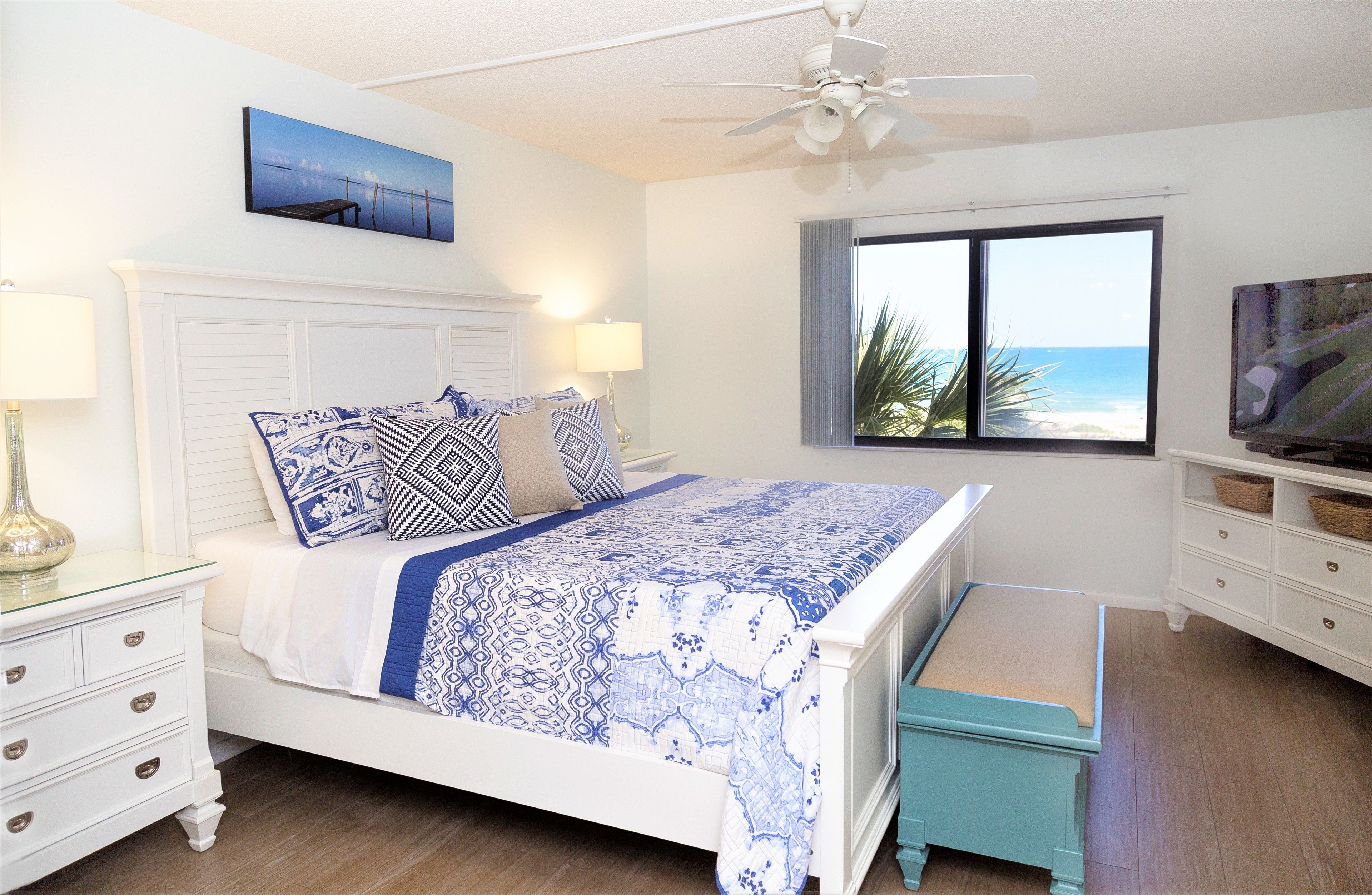 Un dormitorio con cama, mesita de noche, lámpara, televisor, ventana con vista al océano y ventilador de techo.