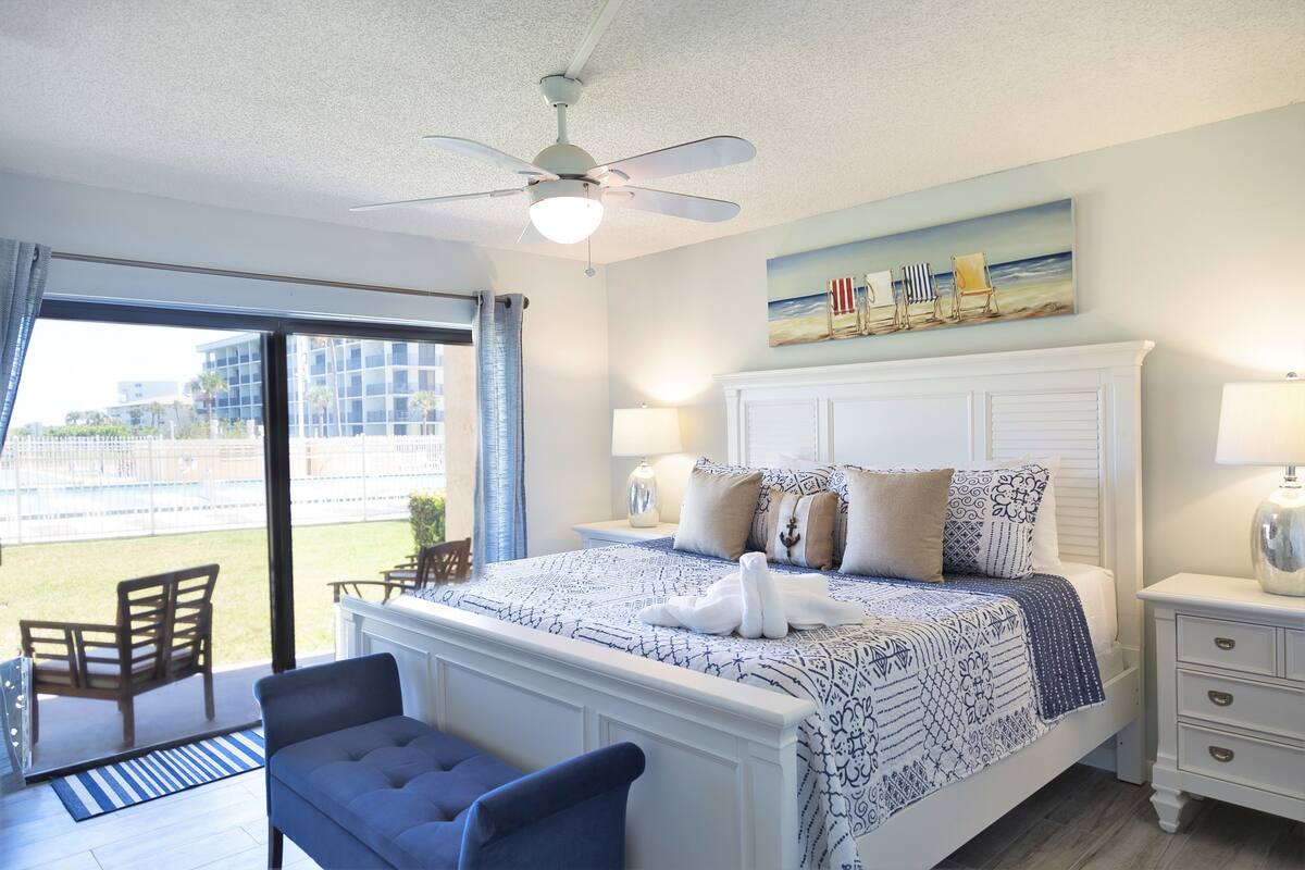 A bedroom with a large bed, a ceiling fan, a painting of beach chairs, and a view of the outdoors.