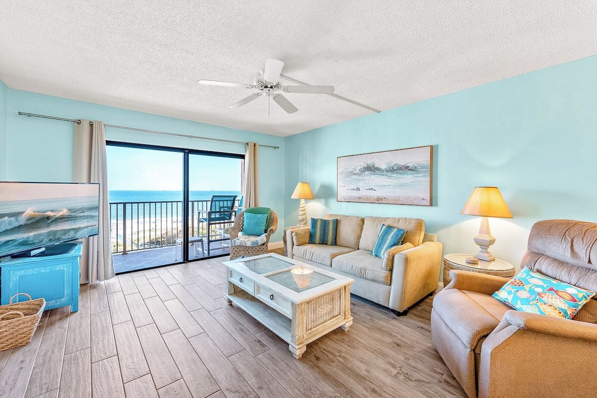 A spacious living room with a large TV, comfortable seating, a ceiling fan, and a balcony with a view of the ocean.