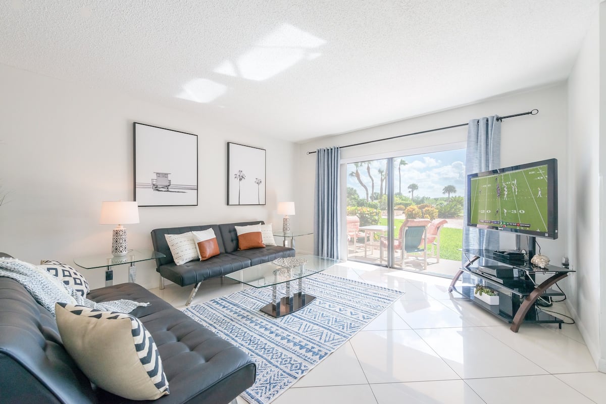 A modern living room with a glass coffee table, a dark sofa with patterned cushions, a flat-screen TV, and a large window with a view of greenery.
