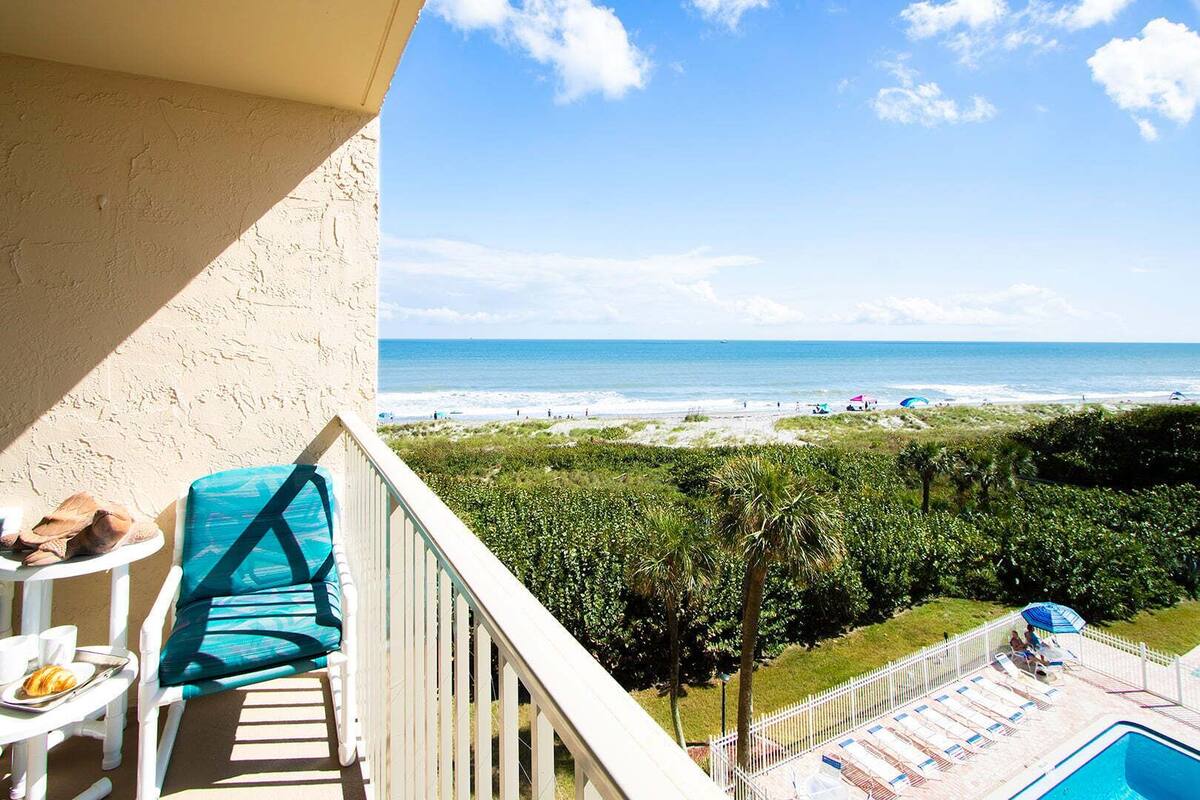 A balcony with a view of the beach, a pool, and a dining area with a croissant.