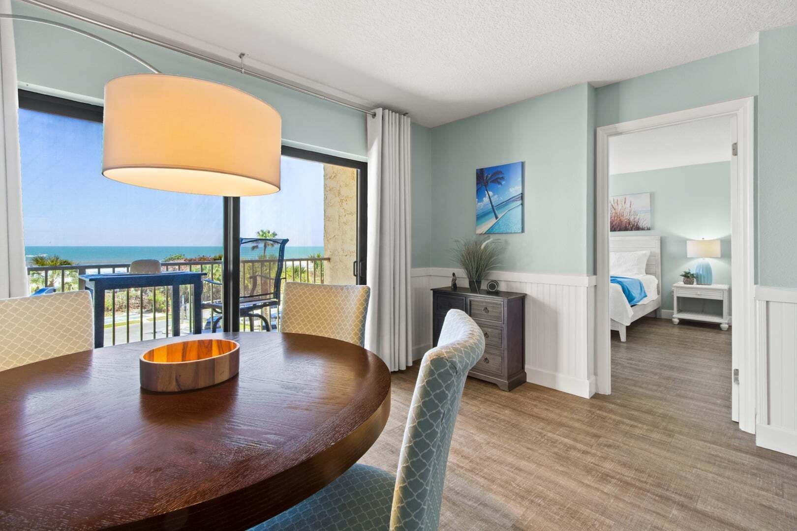Een hotelkamer met een eettafel, stoelen, uitzicht op de oceaan, een bed, een nachtkastje en een schilderij aan de muur.