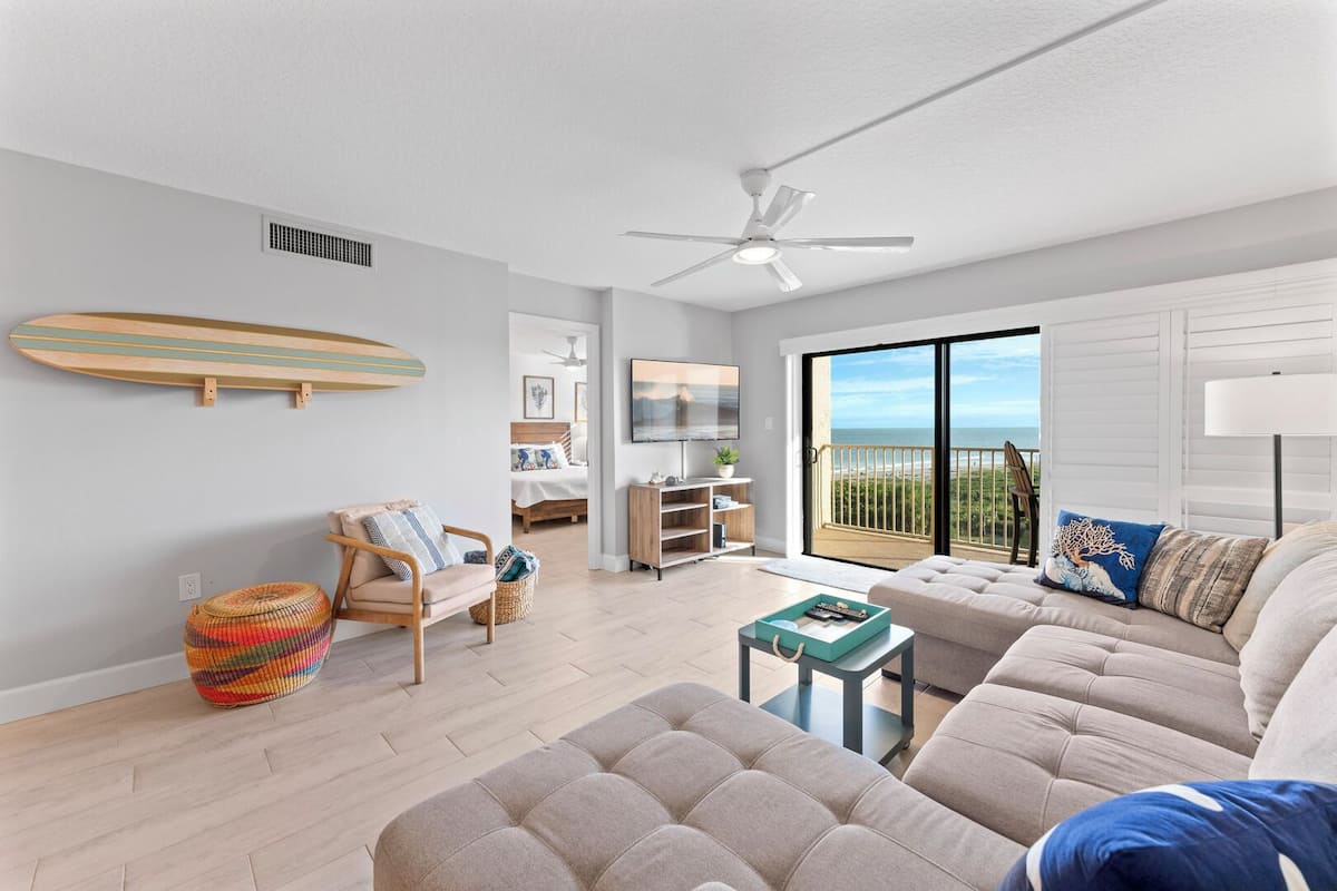A modern living room with a sofa, a chair, a small table, a ceiling fan, and a large window with a view of the ocean.