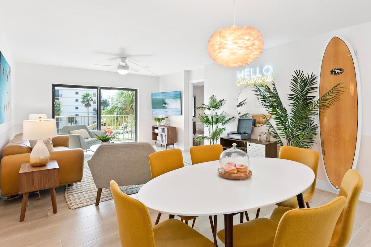 A modern living room with a round dining table, yellow chairs, a ceiling fan, and a surfboard decoration.
