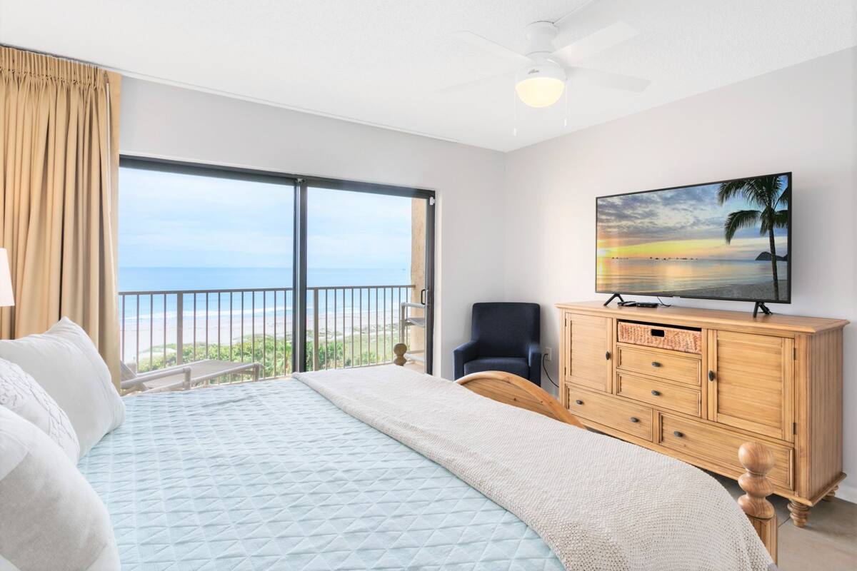 A bedroom with a large bed, a wooden dresser, a television, and a view of the ocean.