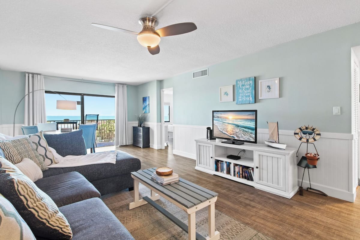 A living room with a sofa, a coffee table, a television, and a balcony with a view of the ocean.