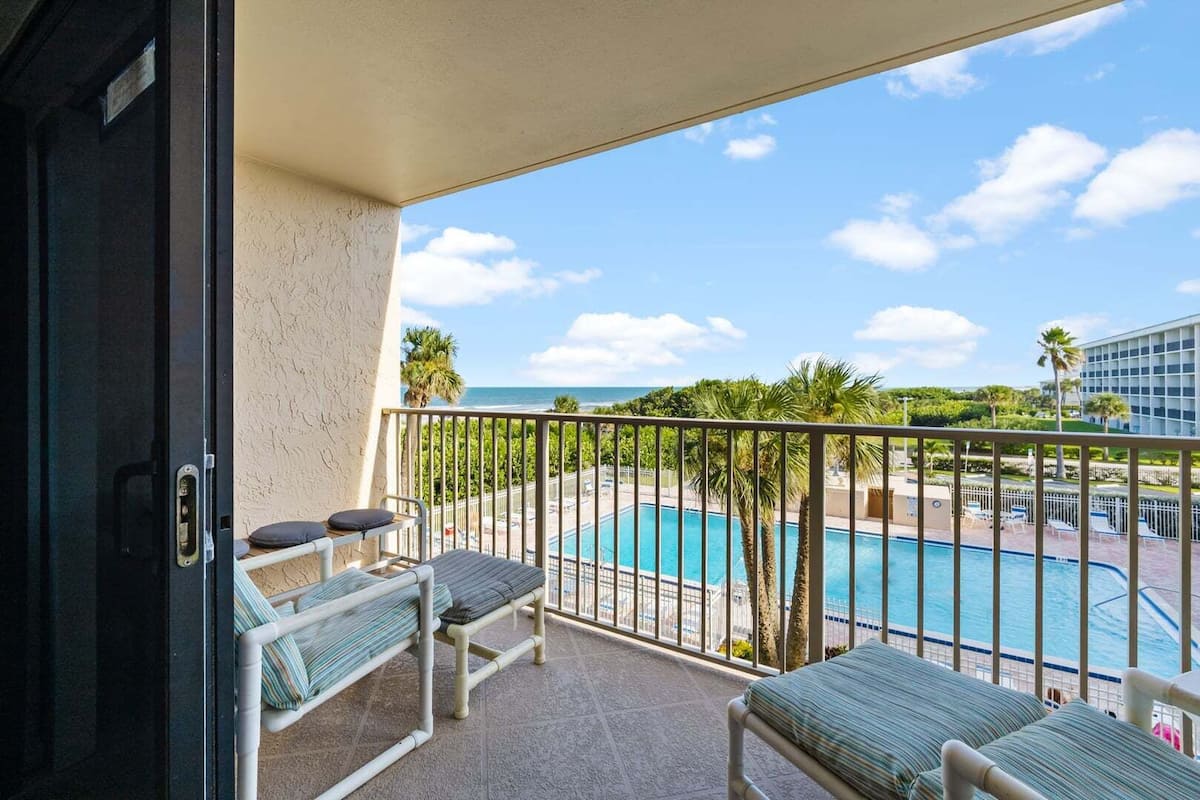 A balcony with two lounge chairs, a pool, and a view of the ocean and greenery.