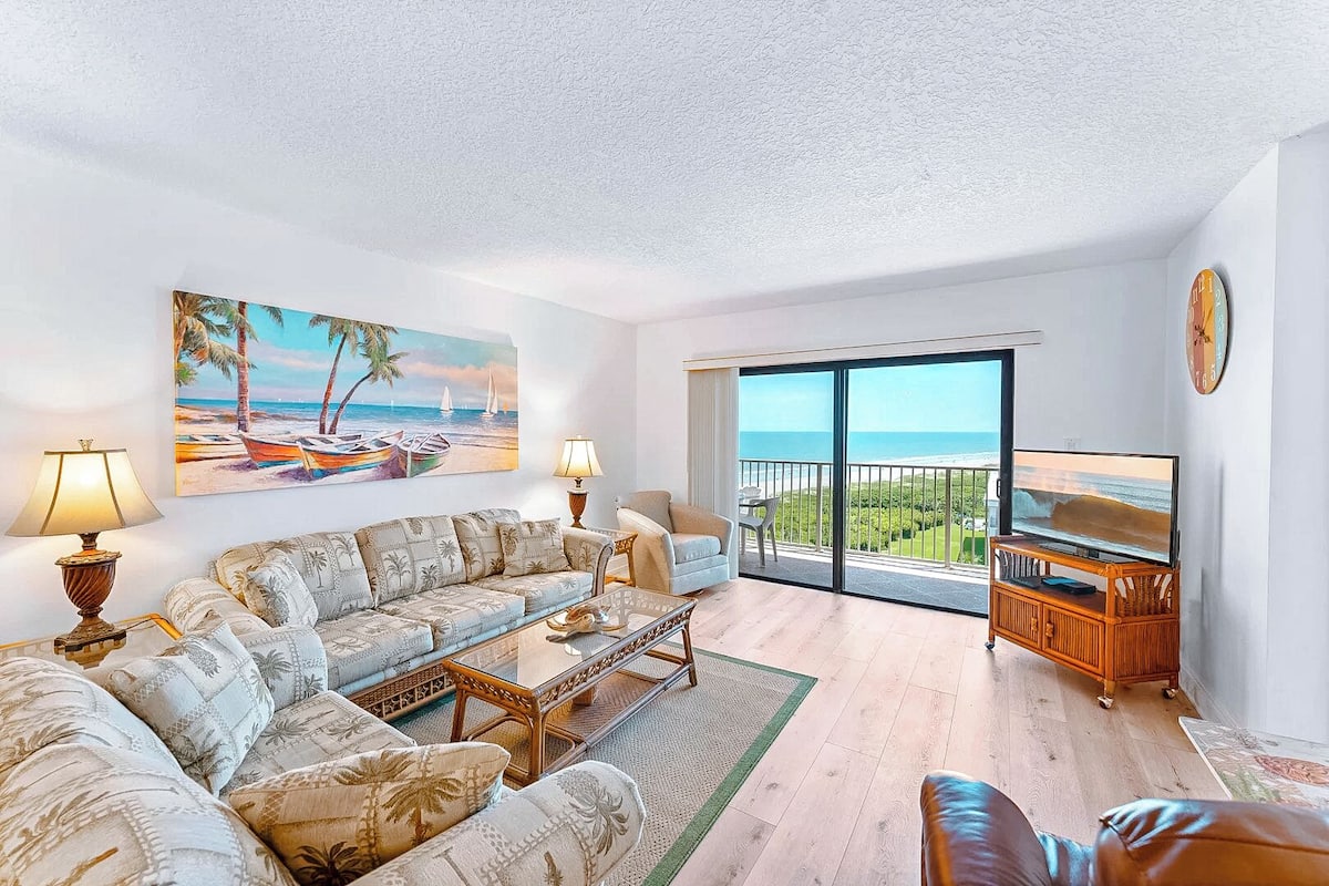 A living room with a sofa, coffee table, and TV, a painting of a beach scene, and a balcony with a view of the ocean.