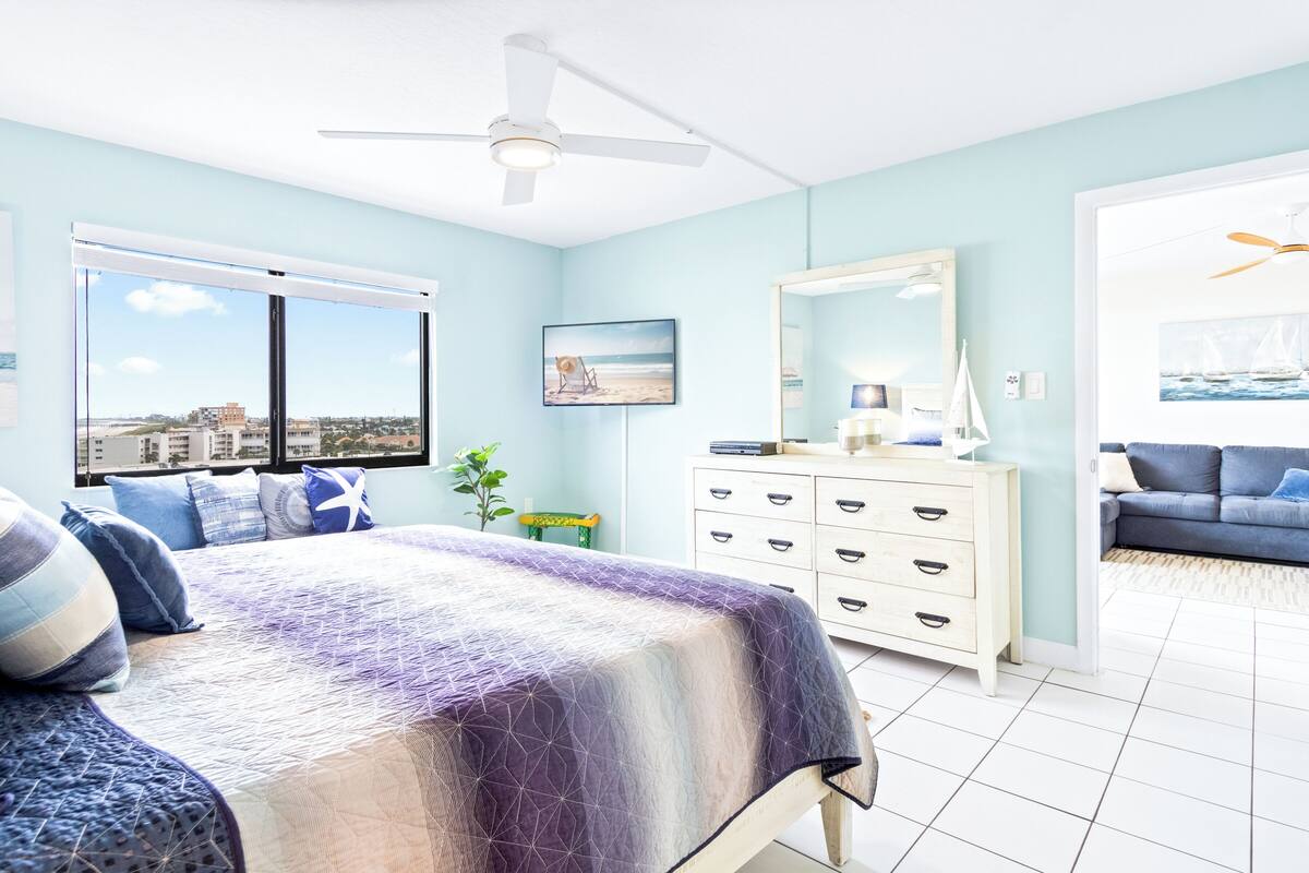 A bedroom with a large bed, a dresser, a mirror, and a view of the city through the window.