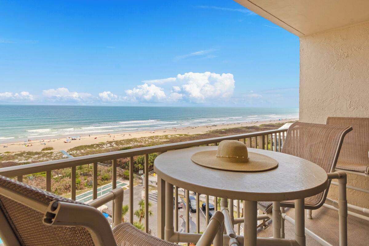 A balcony with a view of the beach, a round table, and a wicker chair.