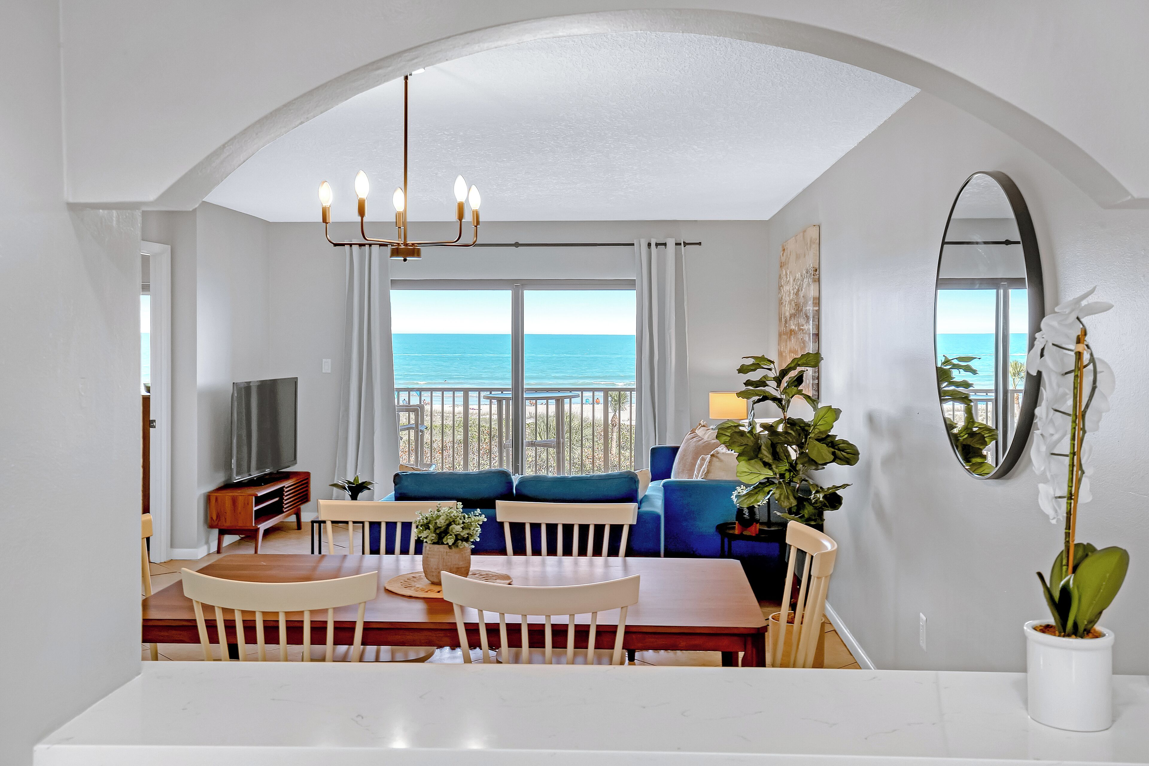 A modern living room with a dining area, large windows with a view of the sea, a chandelier, and a potted plant.