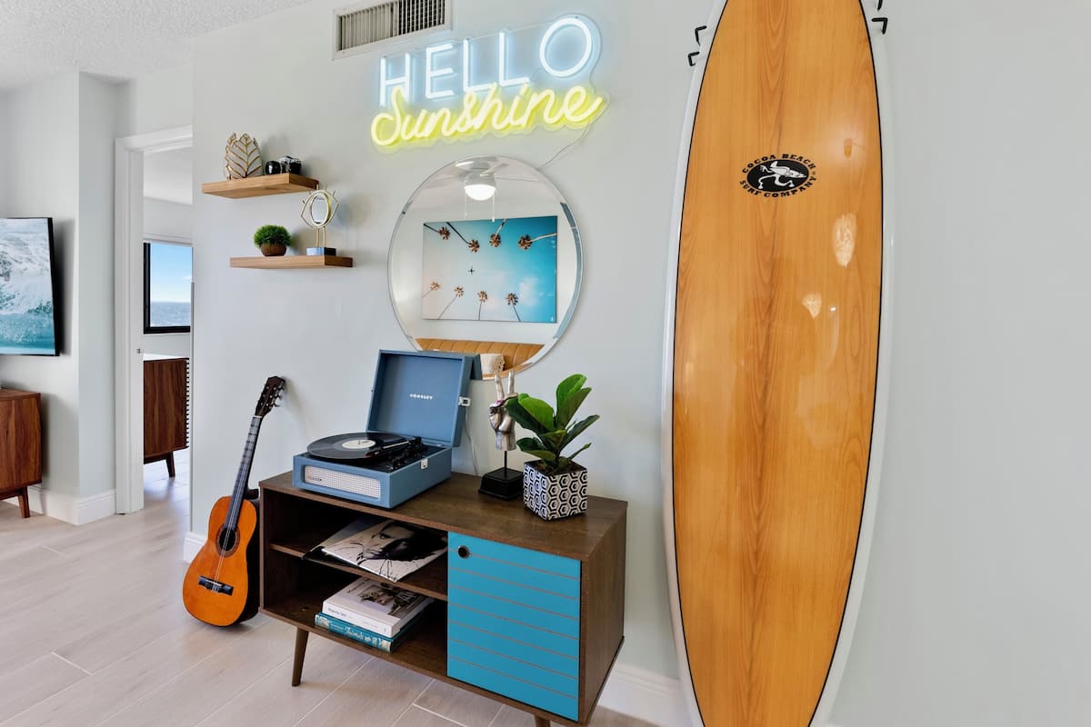 A room with a surfboard, a guitar, a record player, and a neon sign that reads "HELLO Sunshine".