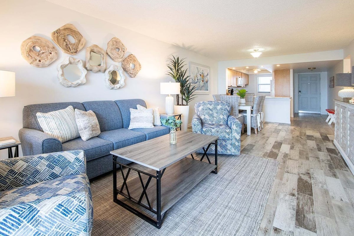 A living room with a sofa, armchair, and coffee table, a kitchen area with cabinets and a dining table, and a hallway leading to a door.
