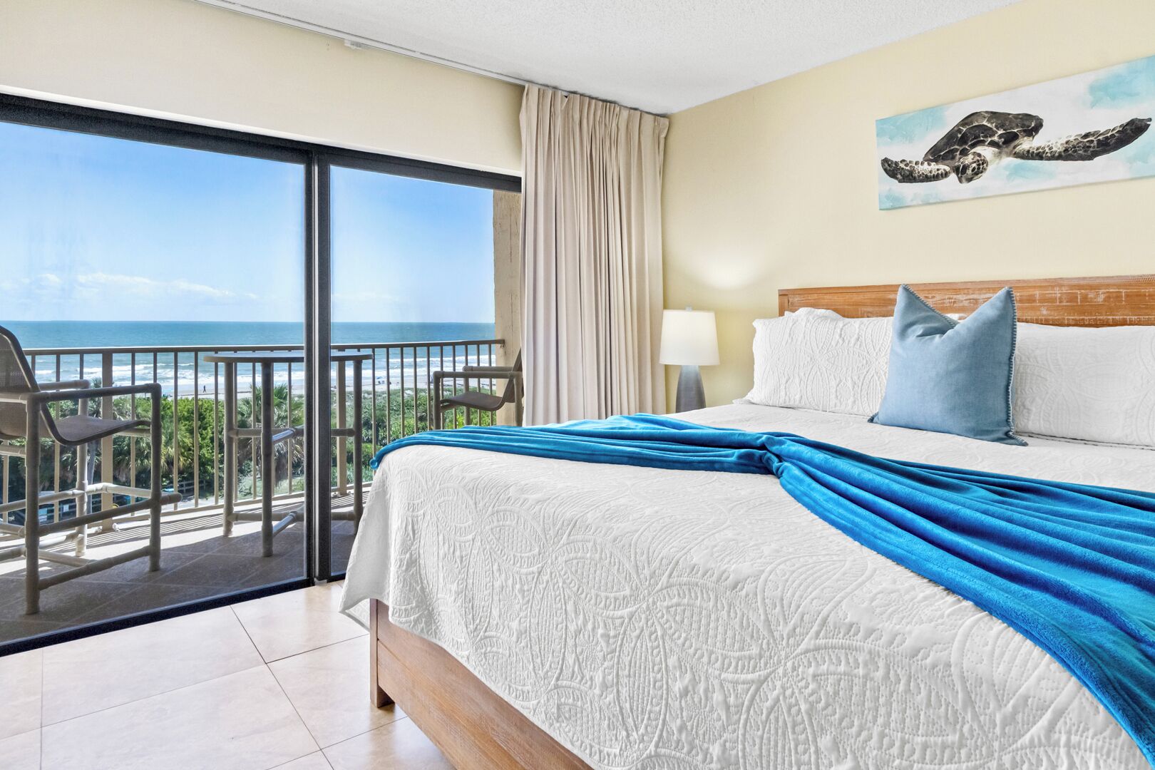 A hotel room with a large bed, a balcony with a view of the ocean, and a painting of a sea turtle on the wall.