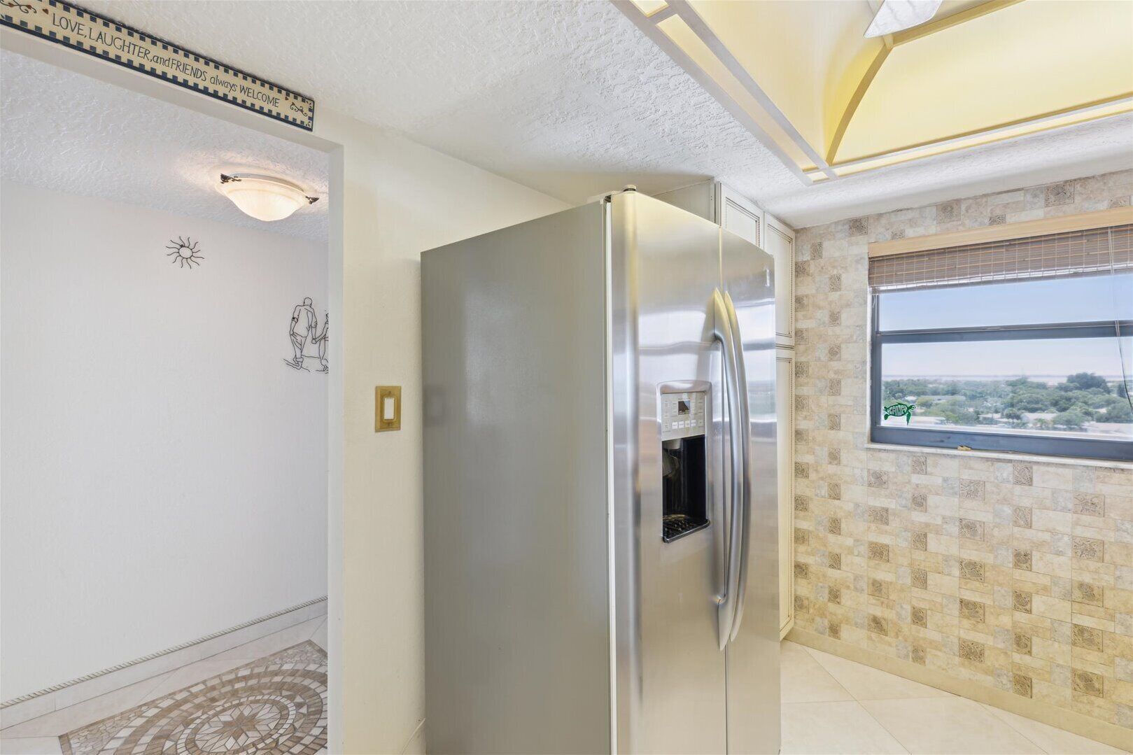 A modern kitchen with a stainless steel refrigerator, a built-in water dispenser, a window with a view, and a decorative wall with a sun and people design.