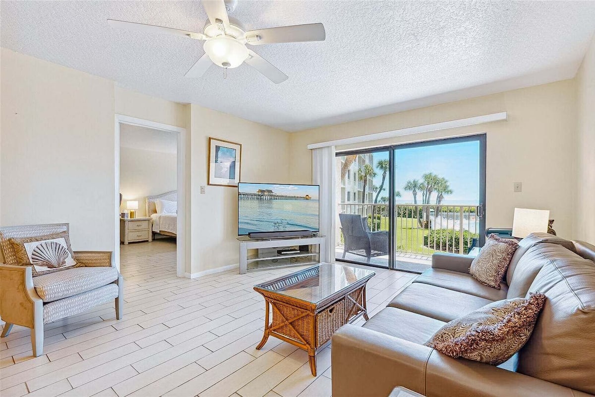 A living room with a television, a ceiling fan, a sofa, a wicker coffee table, and a view of the outdoors.