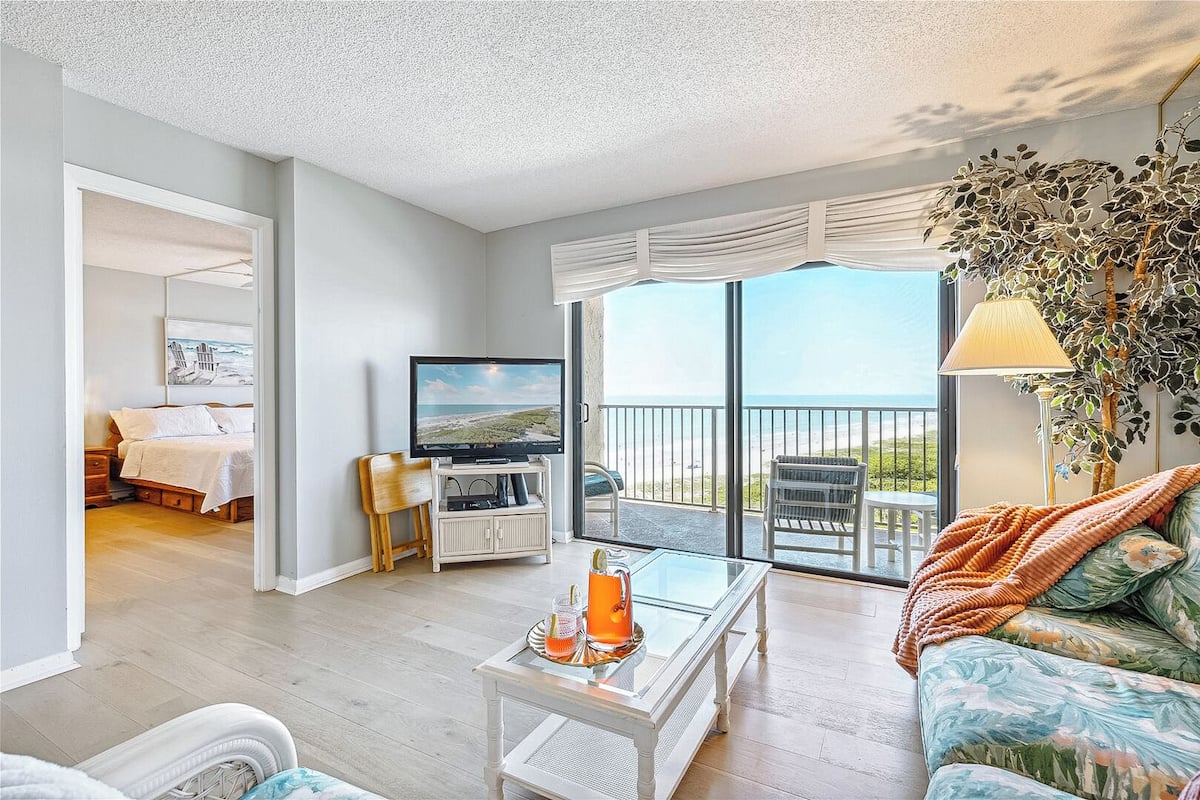 A spacious living area with a balcony, a television, and a view of the beach.
