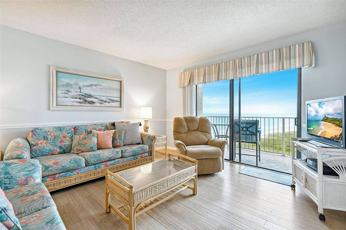 A spacious living room with a sofa, armchair, and a glass coffee table. A large window offers a view of the beach.