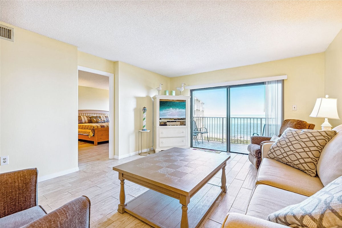 A spacious living room with a wooden coffee table, a sofa, and a view of the ocean through a sliding glass door.