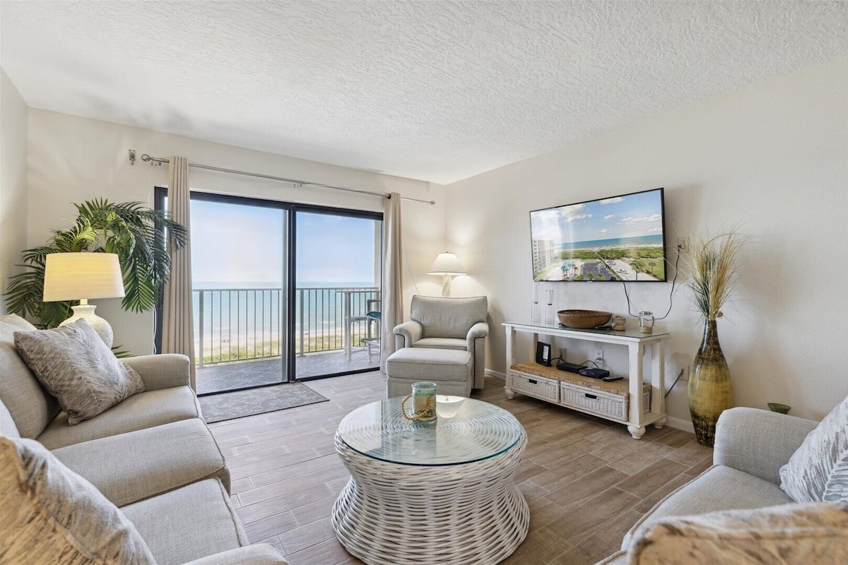 A living room with a sofa, armchair, glass coffee table, TV, and a view of the ocean through a sliding door.