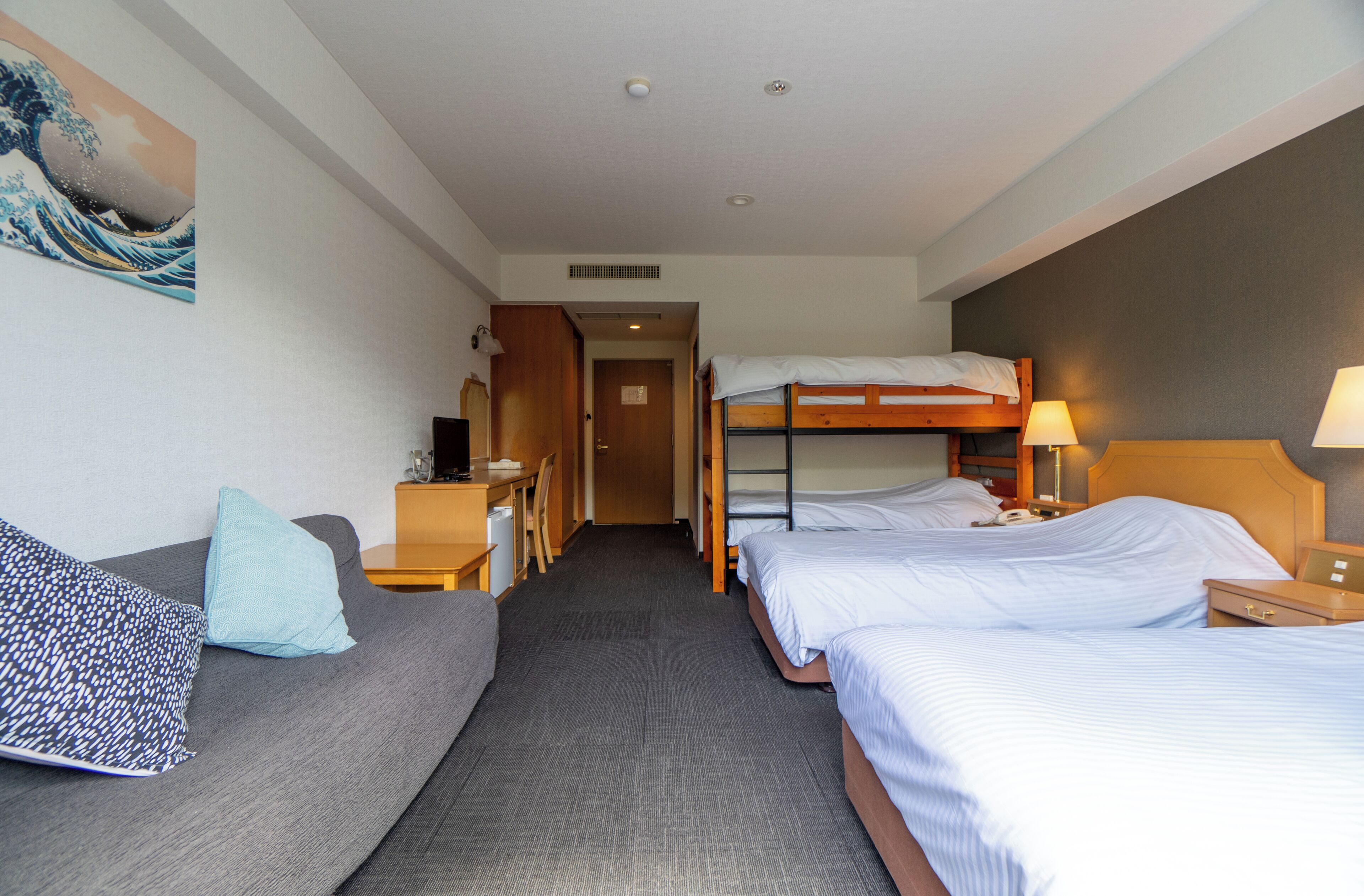 A hotel room with a bunk bed, a sofa, a desk, and a bed with a lamp.