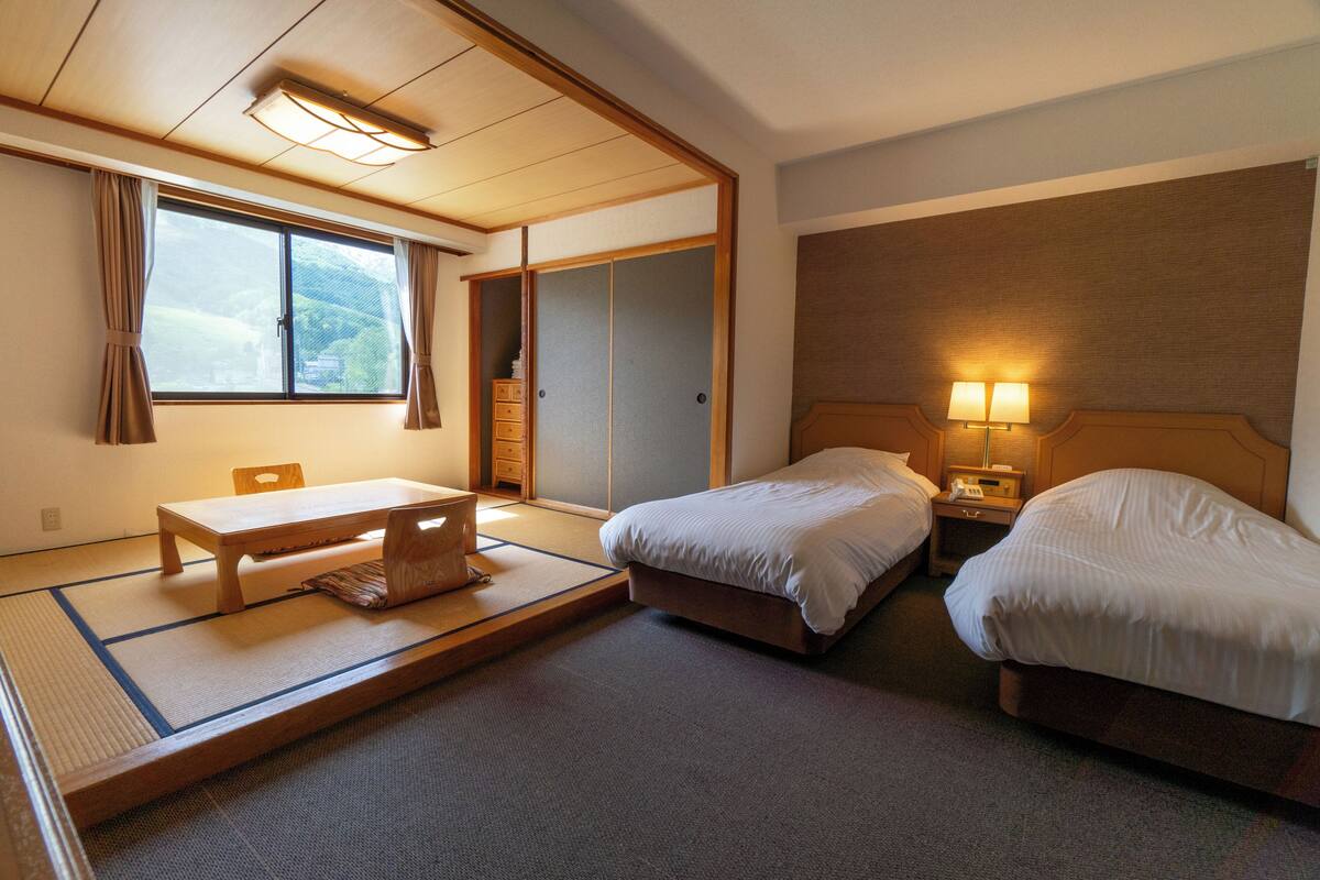 Habitació d'hotel de planta tradicional japonesa amb paviment de tatami, una taula de fusta baixa, dos llits, un armari i una finestra amb cortines.