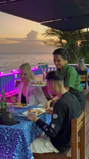 Un comedor con una mesa preparada para tres personas, vista al mar y un atardecer de fondo.