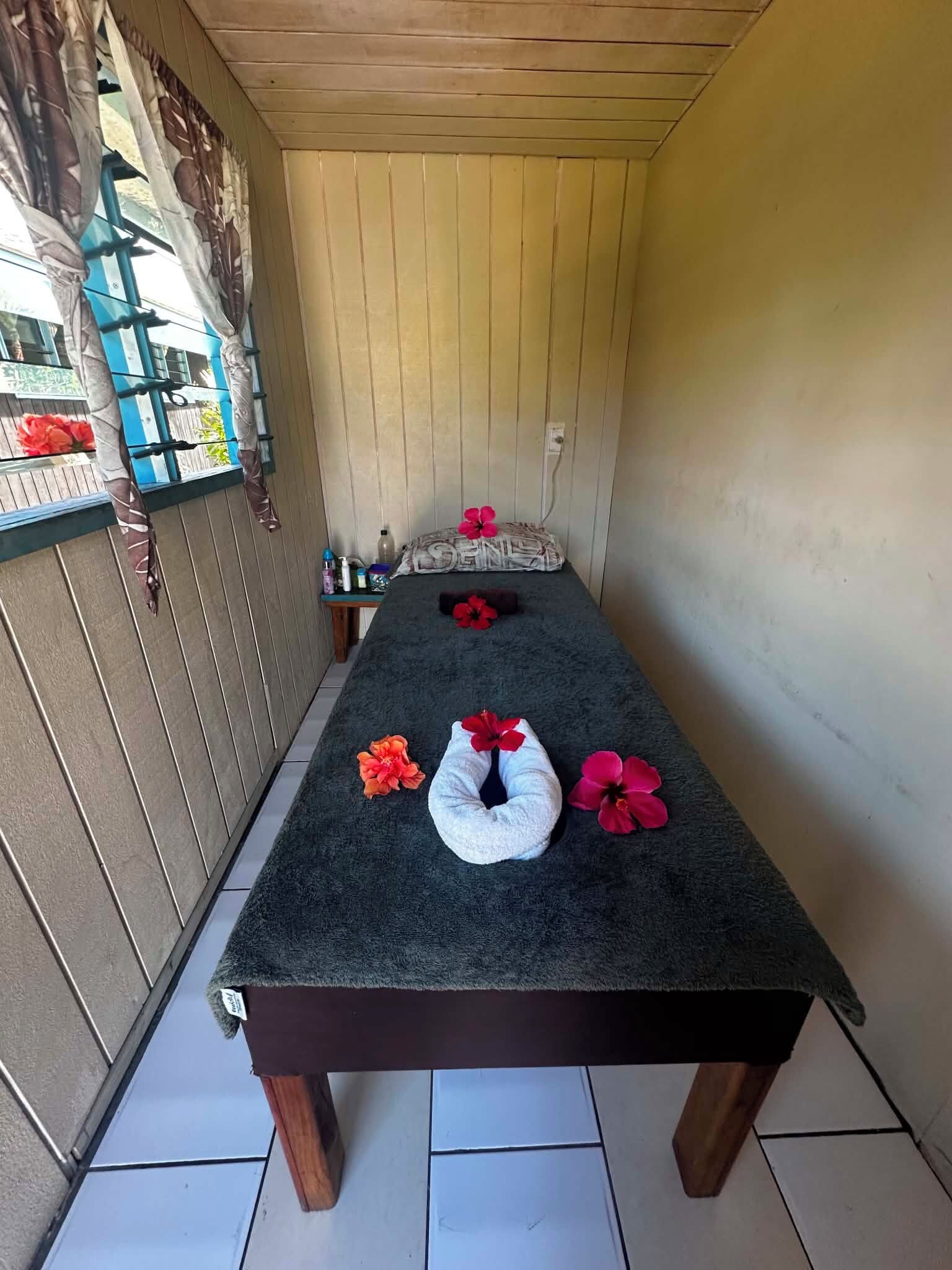 A massage table with towels and flowers, wooden floor, and a window with curtains.