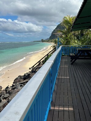 Una terraza de madera con baranda azul que da a una playa de aguas turquesas cristalinas y un litoral rocoso.