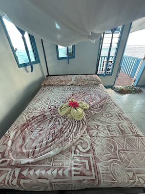 A bed with patterned bedding and a decorative pillow, a window with curtains, and a view of the sea through a balcony.