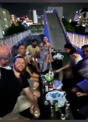 A group of people seated around a table with bottles and glasses, in an outdoor setting with a railing and buildings in the background. - Osaka Namba Hostel MIYABI (Osaka)