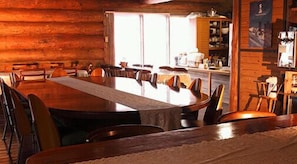 A wooden interior with a long dining table, chairs, and a kitchen area with cabinets and a window. - Canadian Village Goryu (Hakuba)