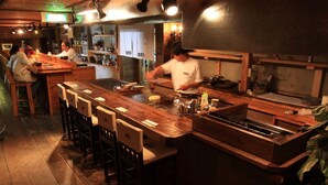 Een restaurant met een lange houten bar, verschillende krukken en een kok die eten bereidt.
