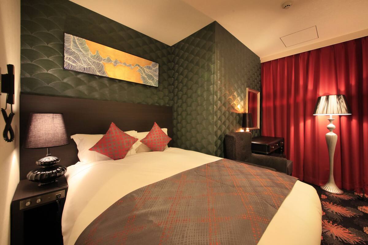 A hotel room with a large bed, a headboard, a nightstand with a lamp, a red curtain, and a wall-mounted artwork.