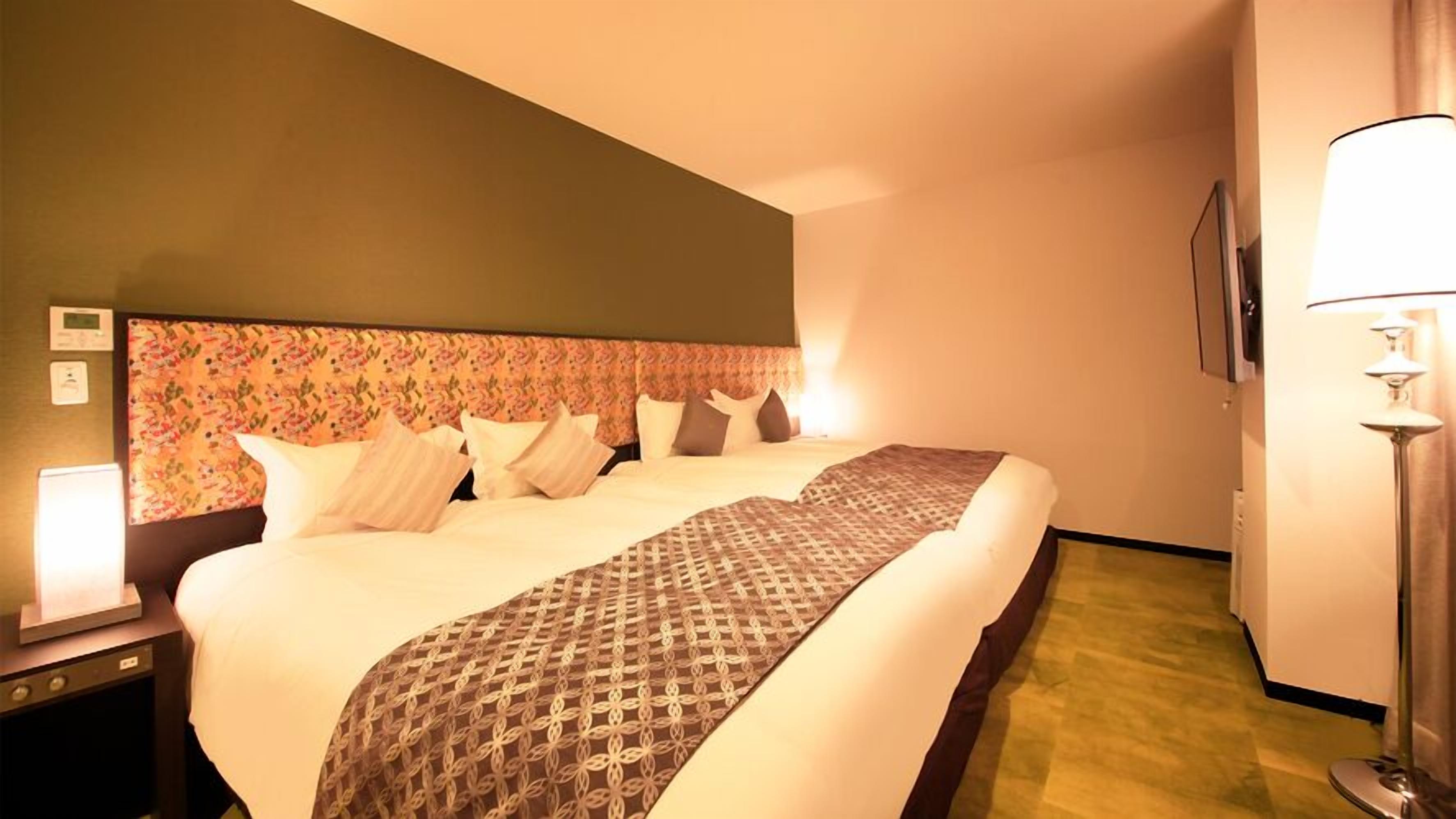 A hotel room with a large bed, a headboard with floral pattern, a bedside table with a lamp, and a wall-mounted TV.