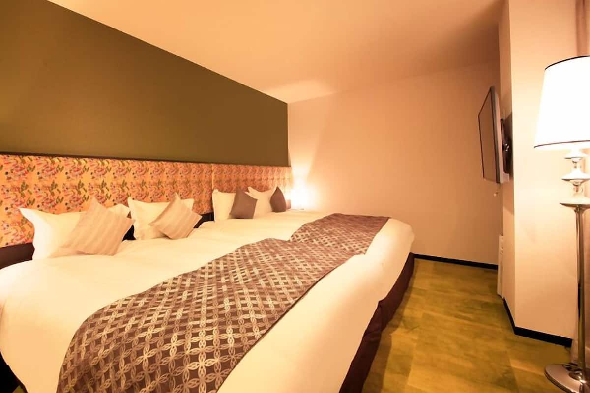 Habitación de hotel con una cama grande, cabecera con estampado floral, mesita de noche con lámpara y televisor montado en la pared.