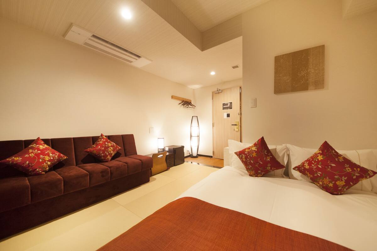 A hotel room with a brown sofa, a bed with white bedding, and a red throw blanket.
