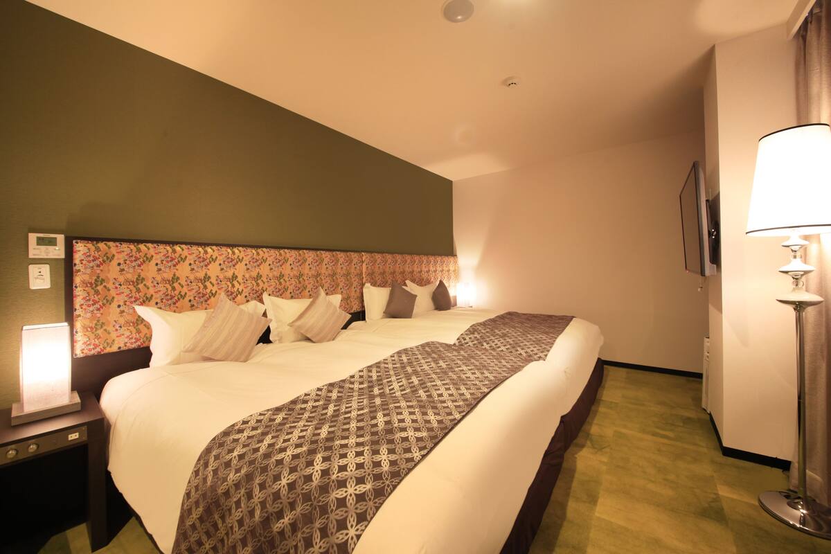 A hotel room with a large bed, a nightstand with a lamp, a wall-mounted TV, and a floor lamp.