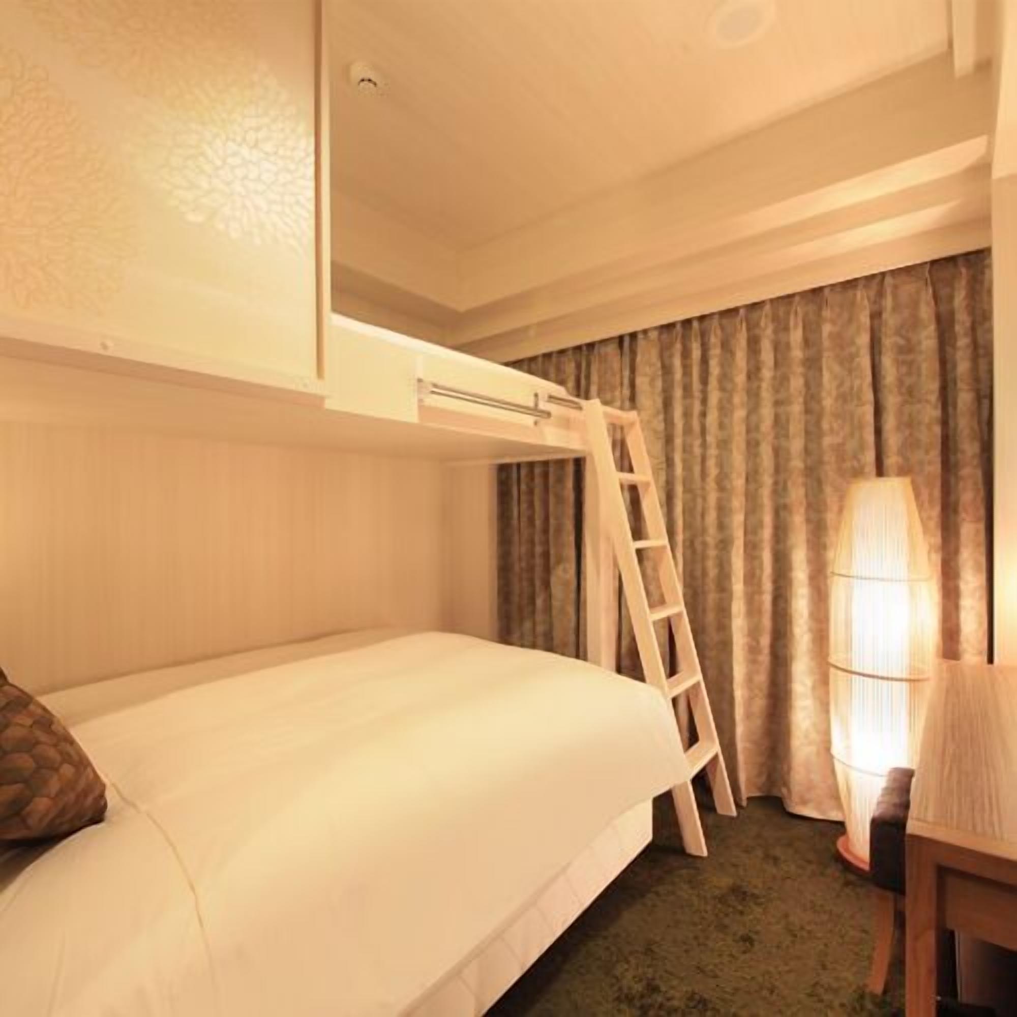 A hotel room with a bunk bed, a desk with a chair, a lamp, and a window with curtains.