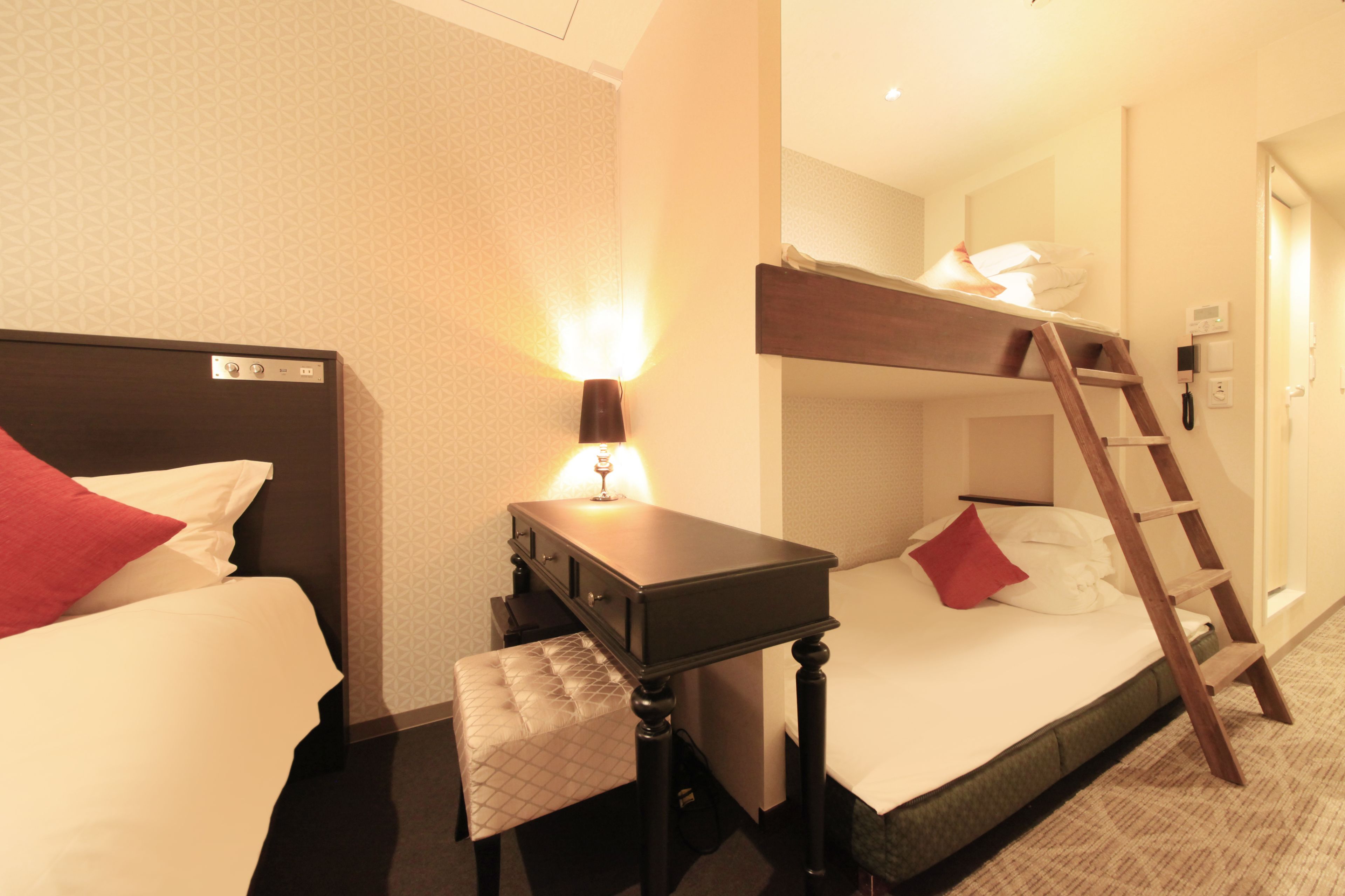 A hotel room with a bunk bed, a bedside table with a lamp, a wooden ladder, and a small bench.