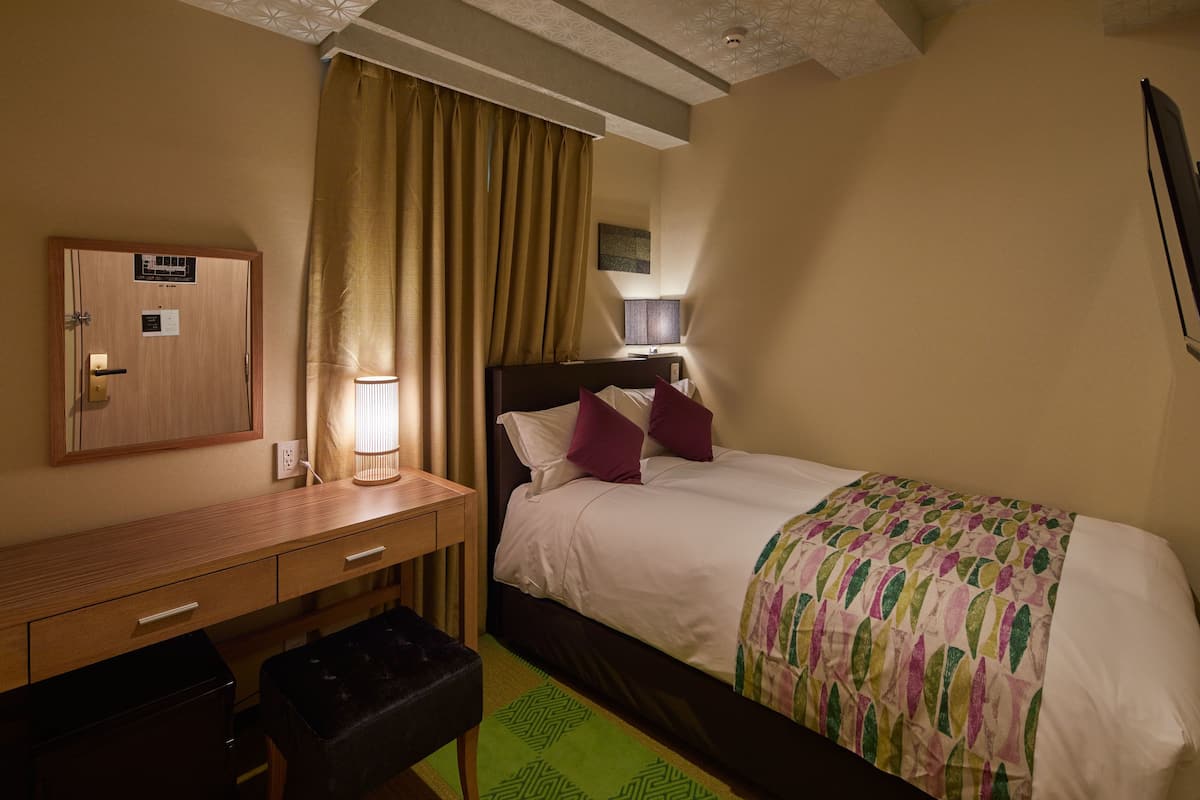 A hotel room with a bed, a desk, a chair, a lamp, and a window with curtains.
