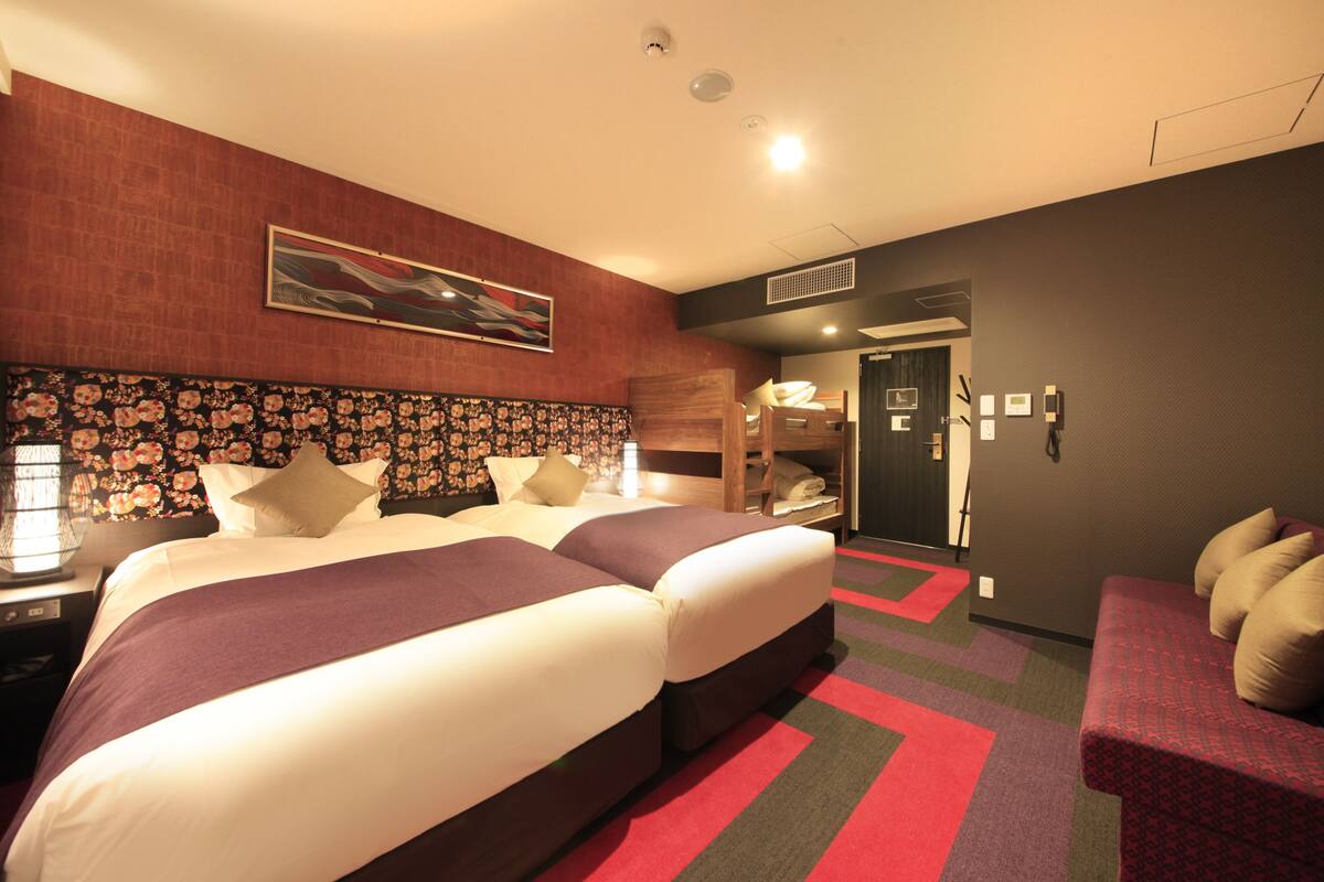 A hotel room with two beds, a sofa, a wooden headboard, a floral-patterned wall, and a red and grey carpet.