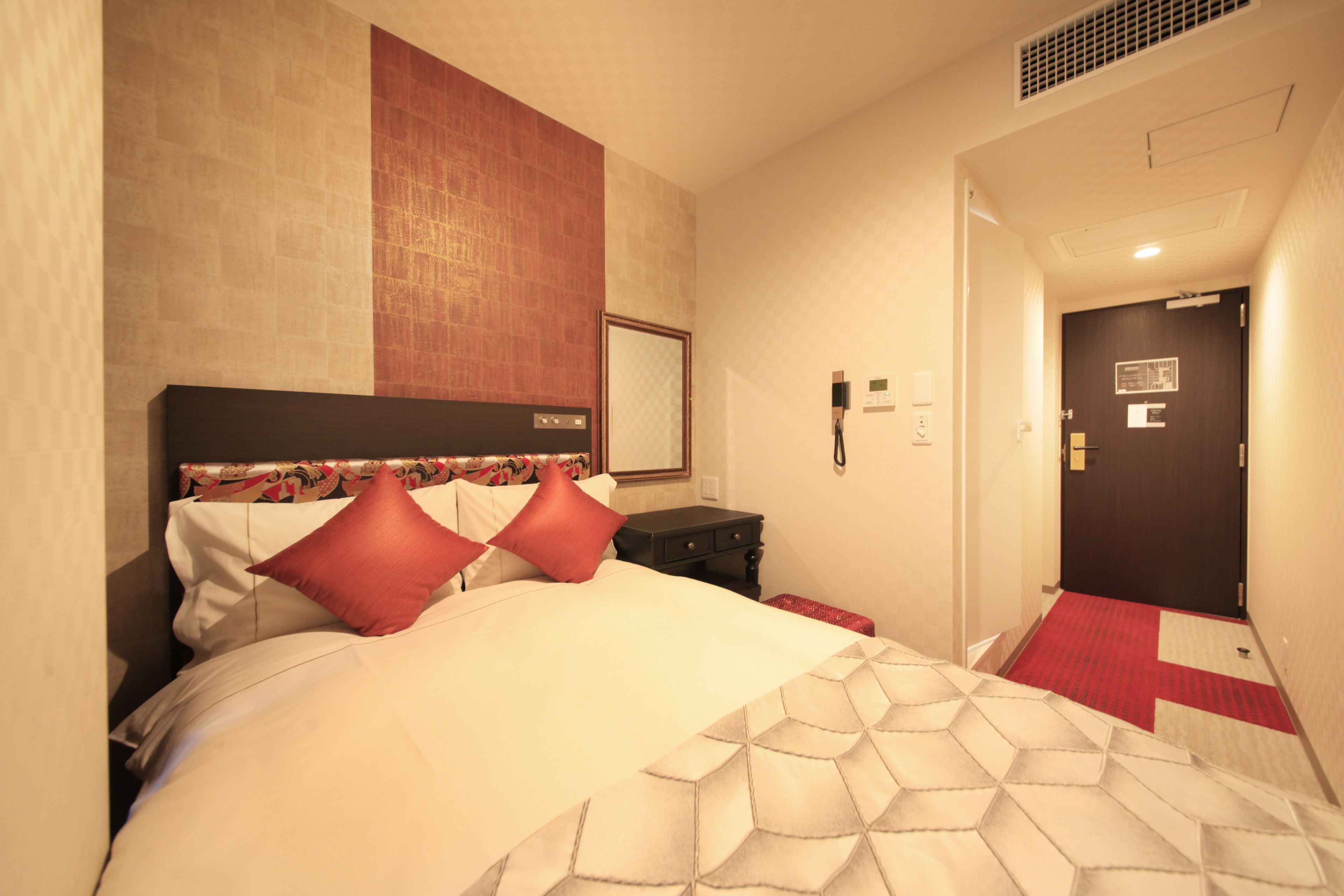 A hotel room with a large bed, a nightstand, a red carpeted hallway, and a door with a sign.