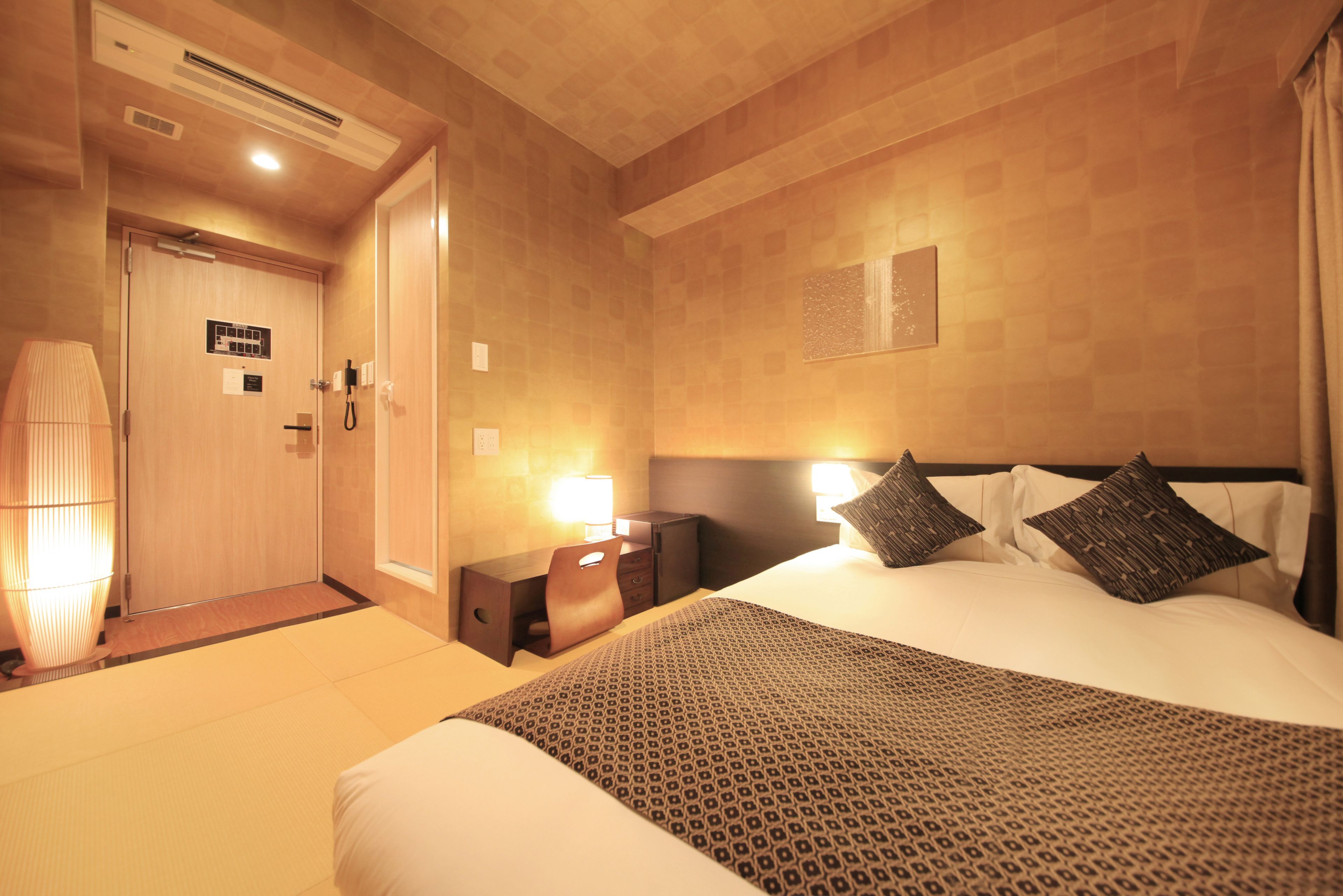 A hotel room with a bed, bedside table, lamp, and a door leading to a bathroom.